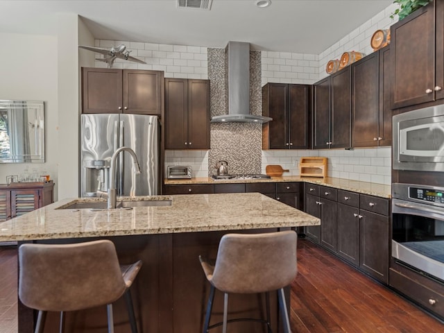 kitchen with wall chimney range hood, an island with sink, dark hardwood / wood-style floors, sink, and stainless steel appliances