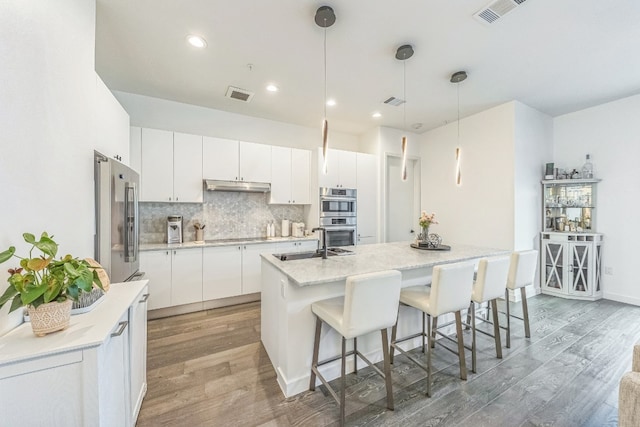 kitchen featuring appliances with stainless steel finishes, an island with sink, white cabinetry, decorative light fixtures, and hardwood / wood-style flooring