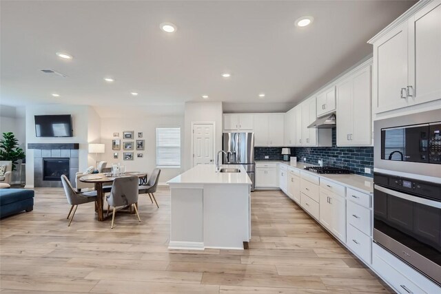 kitchen featuring a tile fireplace, stainless steel appliances, light hardwood / wood-style flooring, a center island with sink, and white cabinets