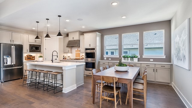 kitchen with appliances with stainless steel finishes, an island with sink, a kitchen breakfast bar, decorative light fixtures, and dark wood-type flooring