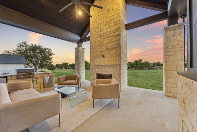 patio terrace at dusk featuring a yard, a grill, an outdoor stone fireplace, exterior kitchen, and ceiling fan