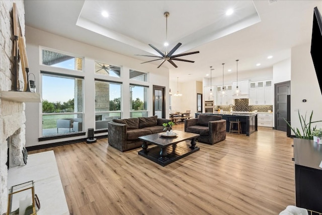 living area with a raised ceiling, ceiling fan with notable chandelier, a stone fireplace, and light wood-style flooring
