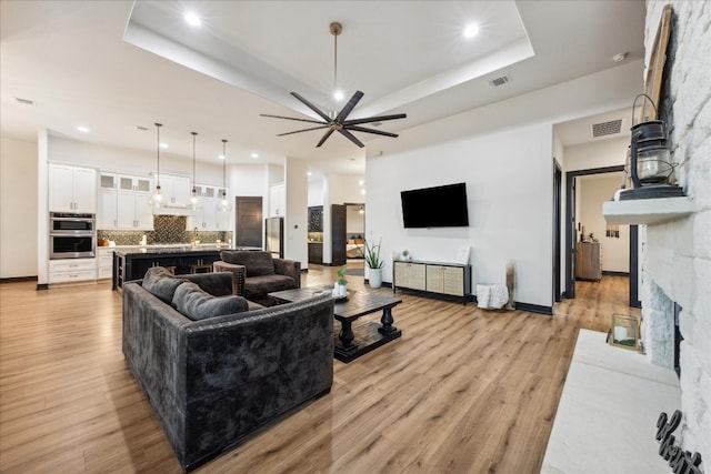 living room featuring light hardwood / wood-style flooring, a stone fireplace, a raised ceiling, and ceiling fan