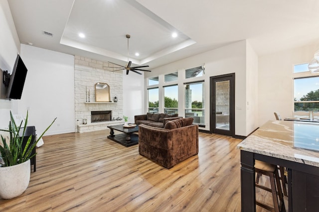 living room featuring recessed lighting, light wood-style flooring, a tray ceiling, and a stone fireplace