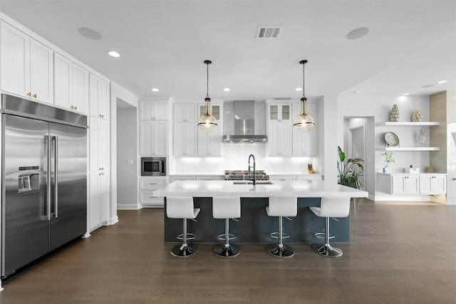 kitchen with wall chimney exhaust hood, hanging light fixtures, built in appliances, an island with sink, and white cabinets