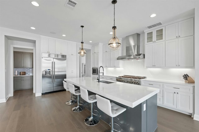 kitchen with sink, wall chimney range hood, built in appliances, an island with sink, and white cabinets