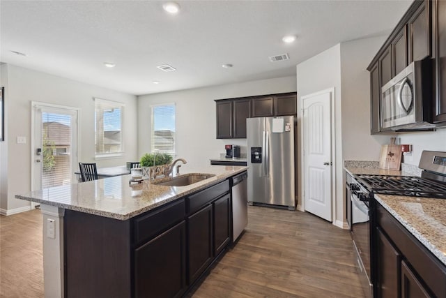 kitchen with appliances with stainless steel finishes, sink, dark hardwood / wood-style flooring, a kitchen island with sink, and light stone countertops