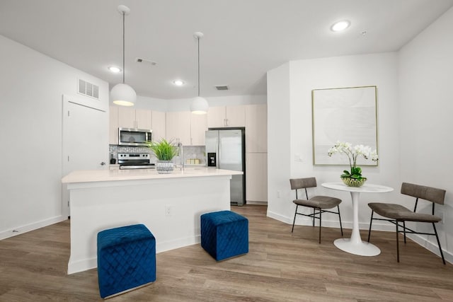 kitchen featuring decorative backsplash, appliances with stainless steel finishes, a kitchen island with sink, pendant lighting, and light hardwood / wood-style flooring