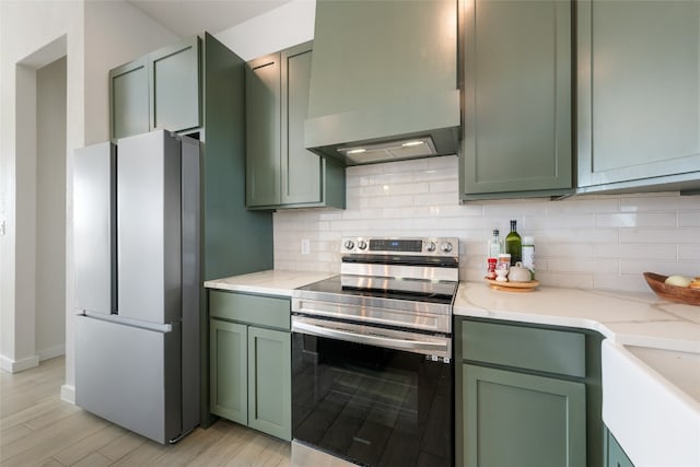 kitchen featuring wall chimney range hood, backsplash, appliances with stainless steel finishes, and green cabinetry