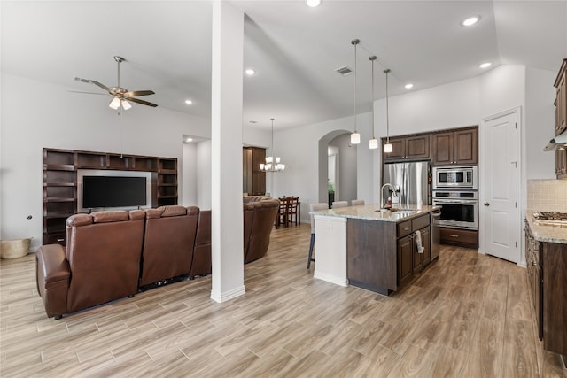 kitchen featuring stainless steel appliances, light stone counters, light hardwood / wood-style flooring, decorative light fixtures, and a center island with sink