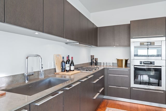 kitchen with sink, light stone counters, light hardwood / wood-style flooring, dark brown cabinetry, and appliances with stainless steel finishes