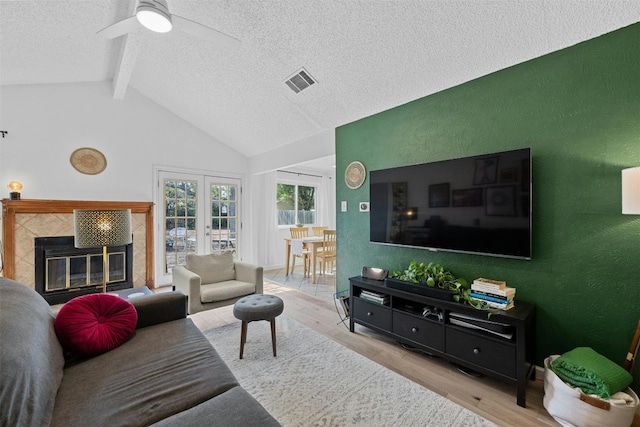 living room featuring vaulted ceiling with beams, french doors, a tiled fireplace, ceiling fan, and light hardwood / wood-style flooring