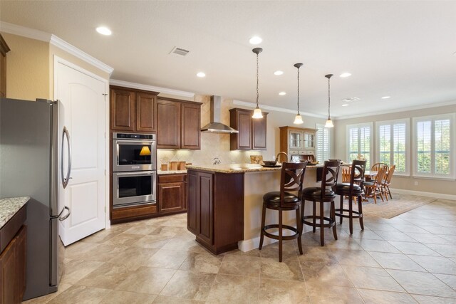 kitchen with an island with sink, appliances with stainless steel finishes, ornamental molding, pendant lighting, and wall chimney exhaust hood