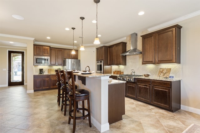 kitchen featuring light stone countertops, wall chimney range hood, an island with sink, hanging light fixtures, and stainless steel appliances