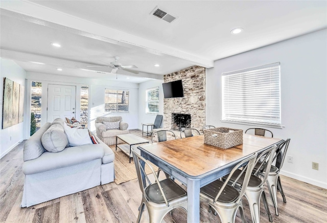 dining room featuring ceiling fan, a stone fireplace, beamed ceiling, and light wood-type flooring