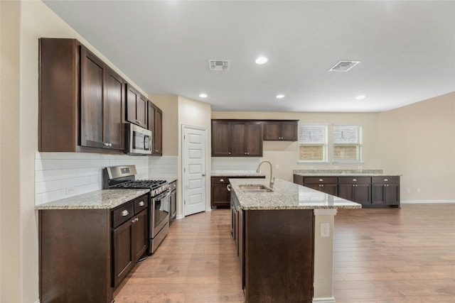 kitchen featuring an island with sink, appliances with stainless steel finishes, light stone countertops, light hardwood / wood-style flooring, and sink