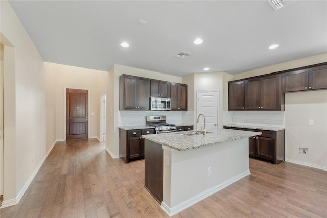 kitchen with light stone counters, appliances with stainless steel finishes, a kitchen island with sink, light wood-type flooring, and sink