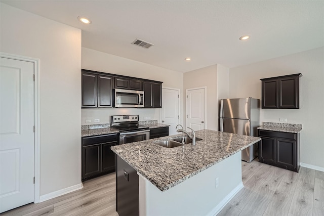 kitchen with light hardwood / wood-style floors, stainless steel appliances, a kitchen island with sink, dark stone countertops, and sink