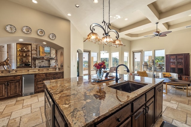 kitchen with hanging light fixtures, a center island with sink, sink, light stone counters, and a towering ceiling