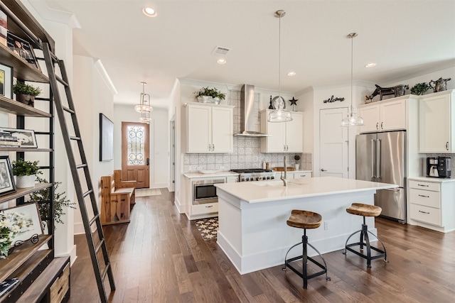 kitchen featuring appliances with stainless steel finishes, wall chimney range hood, a kitchen island with sink, and white cabinets