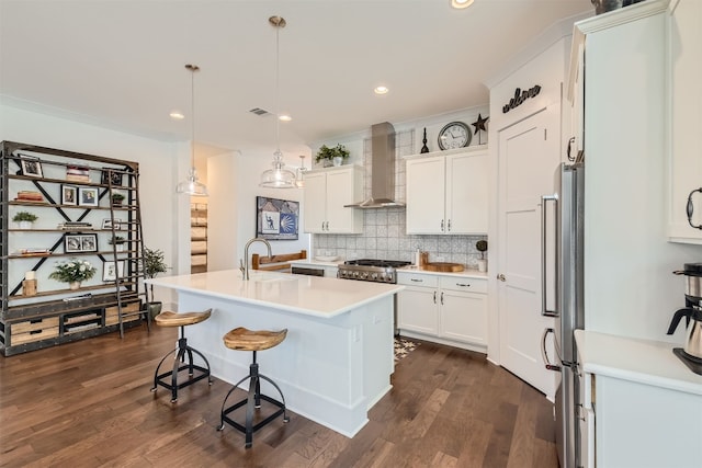 kitchen with wall chimney range hood, white cabinets, an island with sink, appliances with stainless steel finishes, and decorative light fixtures