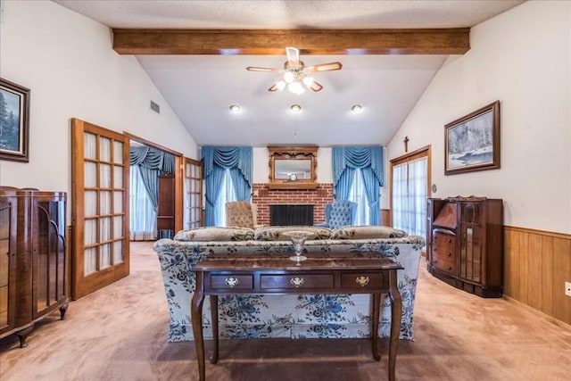 carpeted living room featuring visible vents, lofted ceiling with beams, ceiling fan, wainscoting, and a brick fireplace