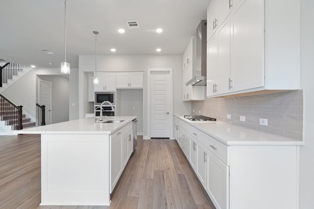 kitchen with wall chimney range hood, an island with sink, stainless steel appliances, light hardwood / wood-style floors, and sink