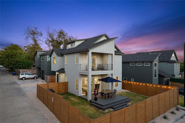 back of property at dusk with driveway, a balcony, a fenced backyard, and board and batten siding