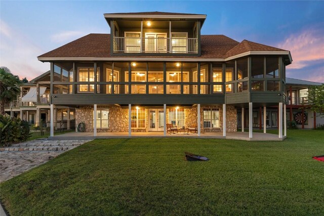 back house at dusk featuring a balcony, a sunroom, a lawn, and a patio