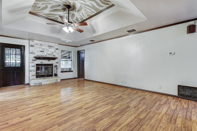 unfurnished living room with ceiling fan, a tray ceiling, light hardwood / wood-style flooring, a stone fireplace, and crown molding