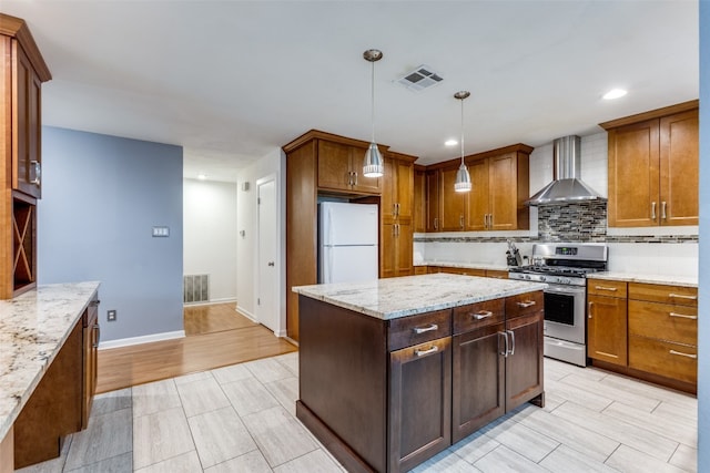 kitchen with wall chimney range hood, white refrigerator, light wood-type flooring, hanging light fixtures, and stainless steel range with gas stovetop