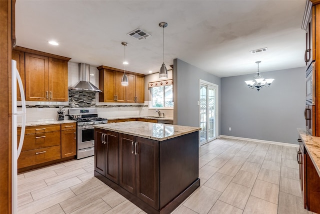 kitchen with stainless steel appliances, wall chimney range hood, light stone countertops, hanging light fixtures, and a center island