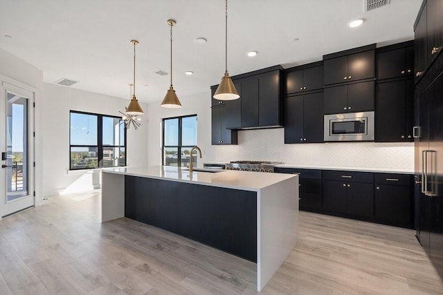 kitchen with stainless steel microwave, a kitchen island with sink, sink, decorative light fixtures, and light hardwood / wood-style floors