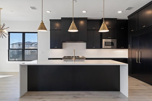 kitchen with built in appliances, a kitchen island with sink, light hardwood / wood-style flooring, and hanging light fixtures