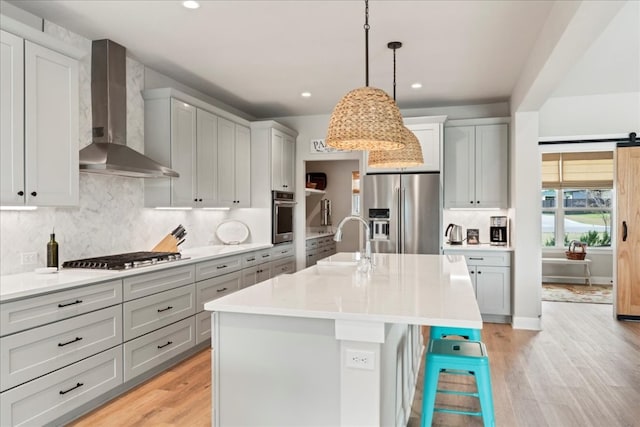 kitchen featuring appliances with stainless steel finishes, wall chimney exhaust hood, light hardwood / wood-style flooring, and a center island with sink