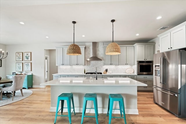 kitchen with wall chimney range hood, a center island with sink, light wood-type flooring, stainless steel appliances, and decorative light fixtures