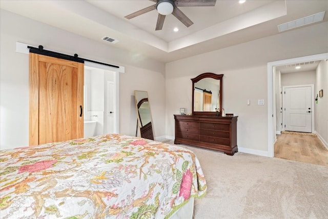 bedroom with a raised ceiling, ceiling fan, a barn door, connected bathroom, and light colored carpet