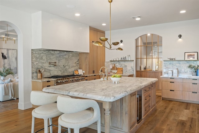 kitchen with light wood-type flooring, backsplash, hanging light fixtures, light stone counters, and a center island with sink