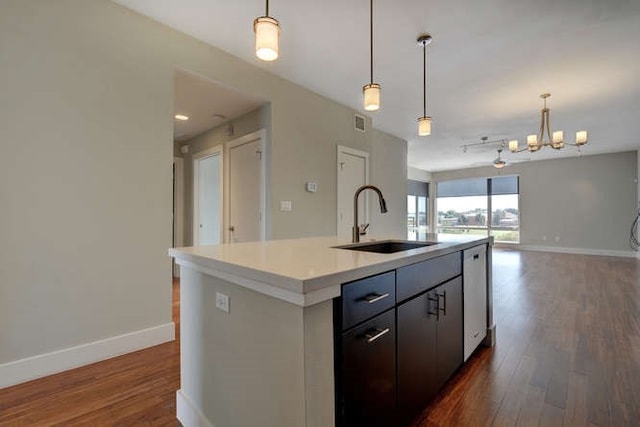 kitchen with sink, hanging light fixtures, a kitchen island with sink, a notable chandelier, and dark wood-type flooring