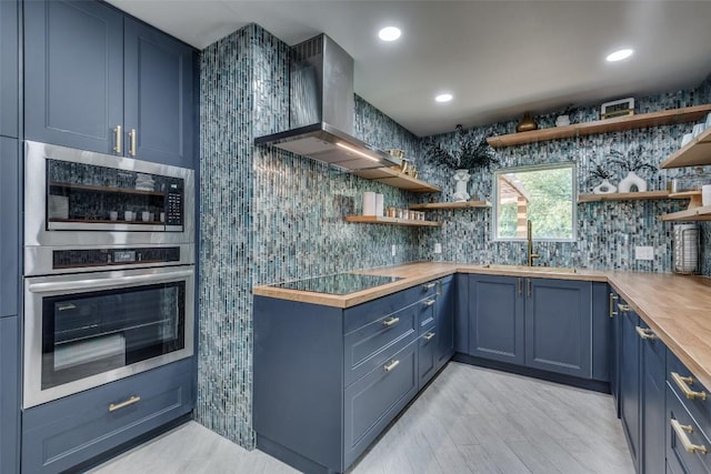 kitchen with blue cabinetry, wall chimney exhaust hood, butcher block counters, sink, and stainless steel oven