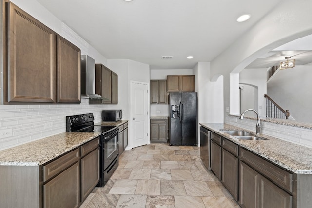 kitchen with black appliances, dark brown cabinets, wall chimney exhaust hood, light stone counters, and sink