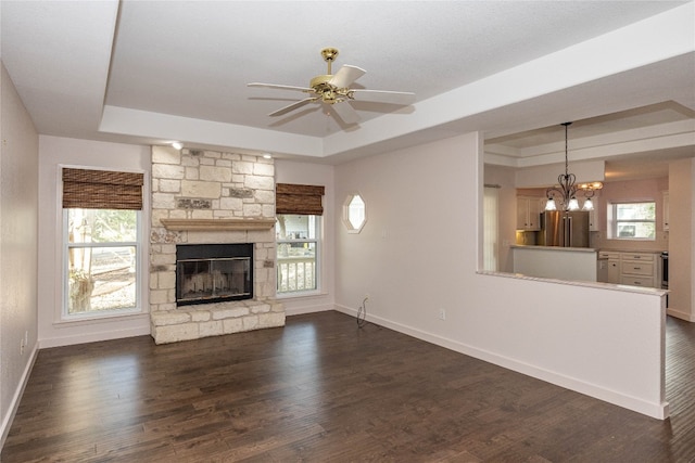 unfurnished living room with dark hardwood / wood-style flooring, a tray ceiling, ceiling fan with notable chandelier, and a stone fireplace