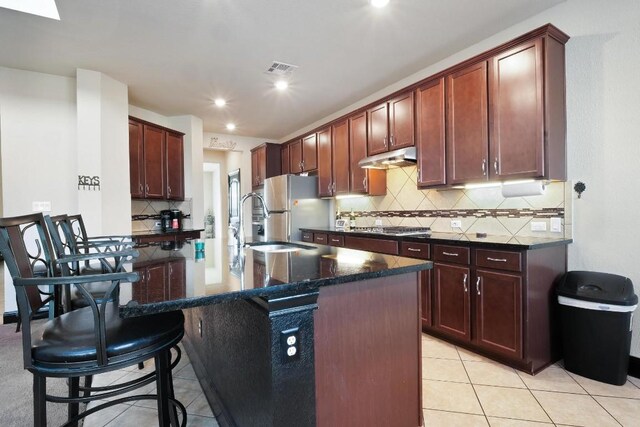 kitchen featuring decorative backsplash, light tile patterned floors, appliances with stainless steel finishes, an island with sink, and dark stone countertops