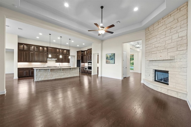 unfurnished living room with sink, a stone fireplace, a tray ceiling, ceiling fan, and dark wood-type flooring