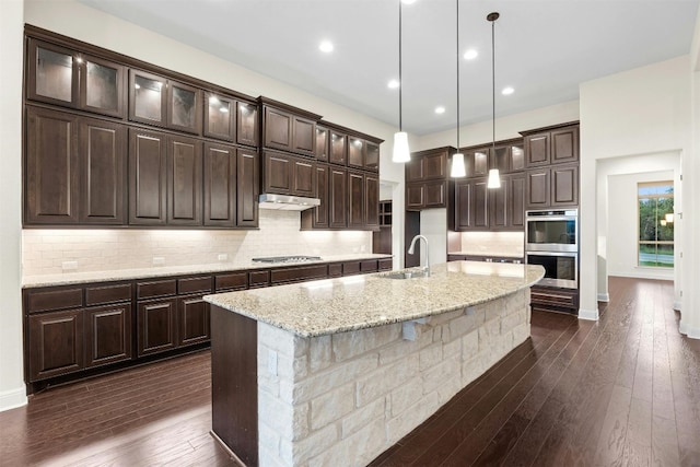 kitchen featuring dark hardwood / wood-style floors, an island with sink, decorative light fixtures, light stone counters, and tasteful backsplash