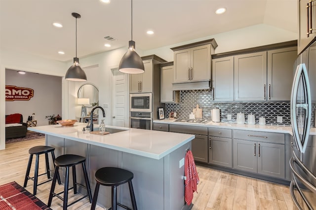 kitchen with gray cabinetry, stainless steel appliances, hanging light fixtures, and a kitchen island with sink