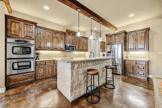 kitchen featuring beam ceiling, light stone counters, backsplash, an island with sink, and appliances with stainless steel finishes