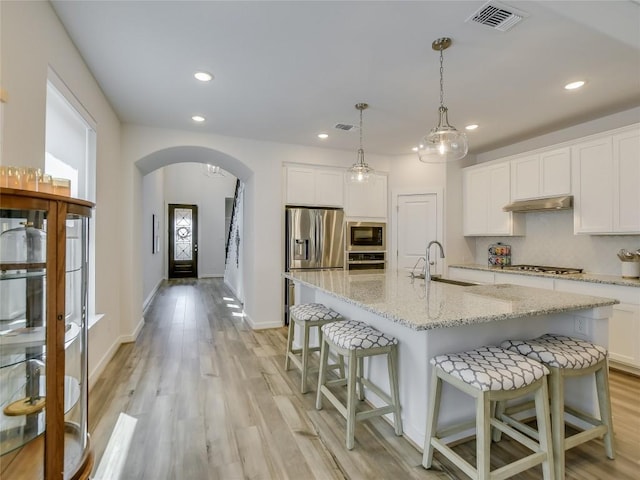 kitchen with appliances with stainless steel finishes, a kitchen island with sink, sink, white cabinets, and hanging light fixtures