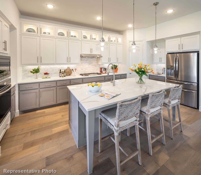 kitchen featuring appliances with stainless steel finishes, an island with sink, hanging light fixtures, light stone counters, and light hardwood / wood-style flooring