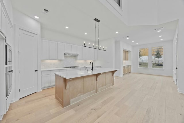 kitchen with an island with sink, oven, white cabinets, gas cooktop, and pendant lighting